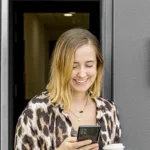 Jasmin lächelt im Türrahmen mit Kaffee und Smartphone.