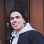 Leon lächelt im Beanie vor einer Holztür.